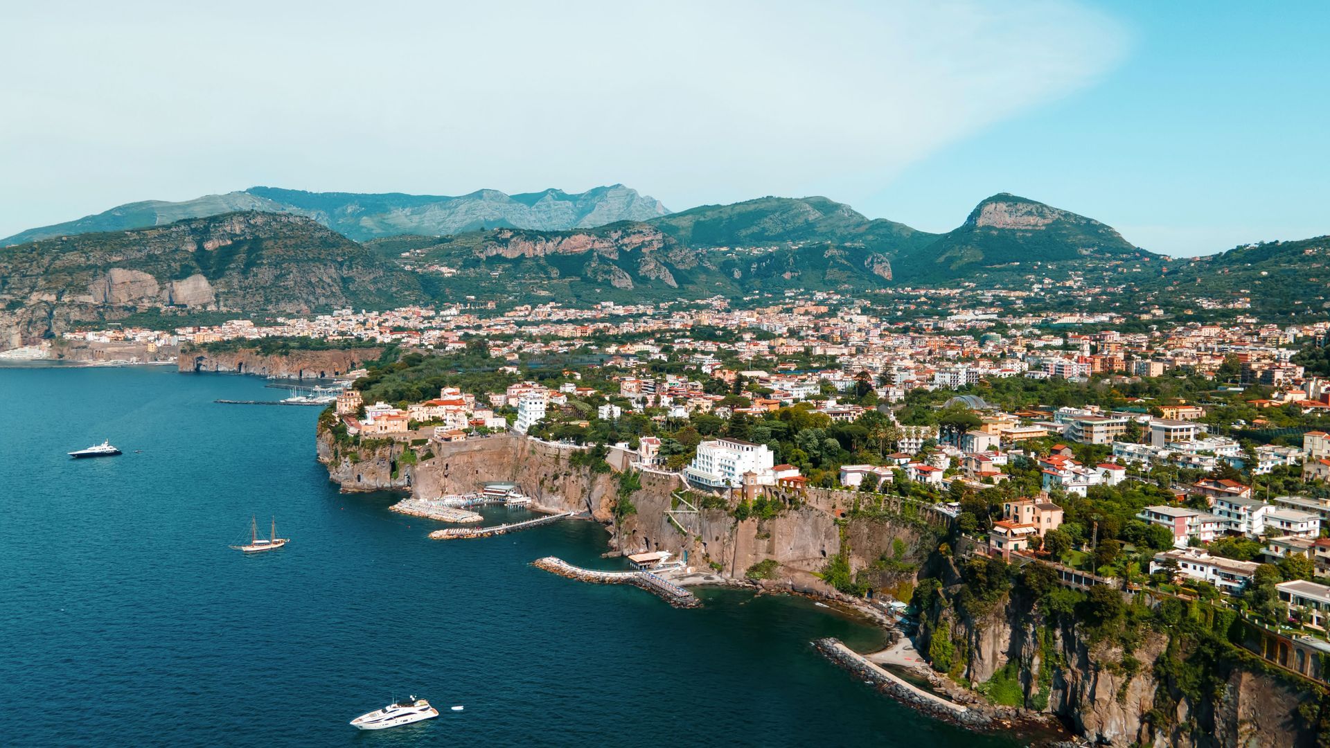 A town perched on high, rugged cliffs overlooking a deep blue sea with boats in the water under a clear sky.