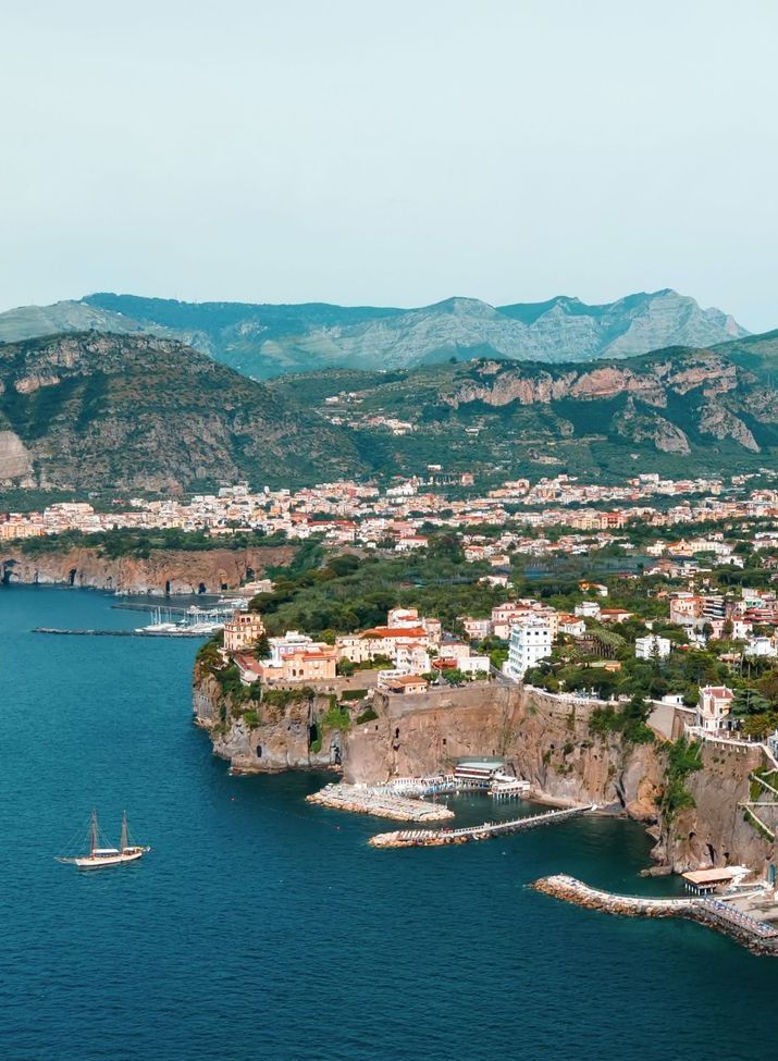 A coastal Italian town with buildings perched on rocky cliffs overlooking a deep blue sea with a sailboat, under mountains.