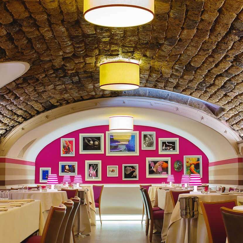 Un ristorante accogliente con soffitto a volta in pietra, tavoli con tovaglie bianche e una parete di fondo di un vivace color magenta.