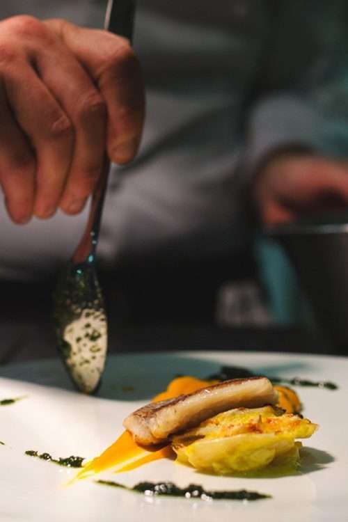 Uno chef usa un cucchiaio per versare con cura la salsa verde su un piatto con pesce scottato e verdure dorate.