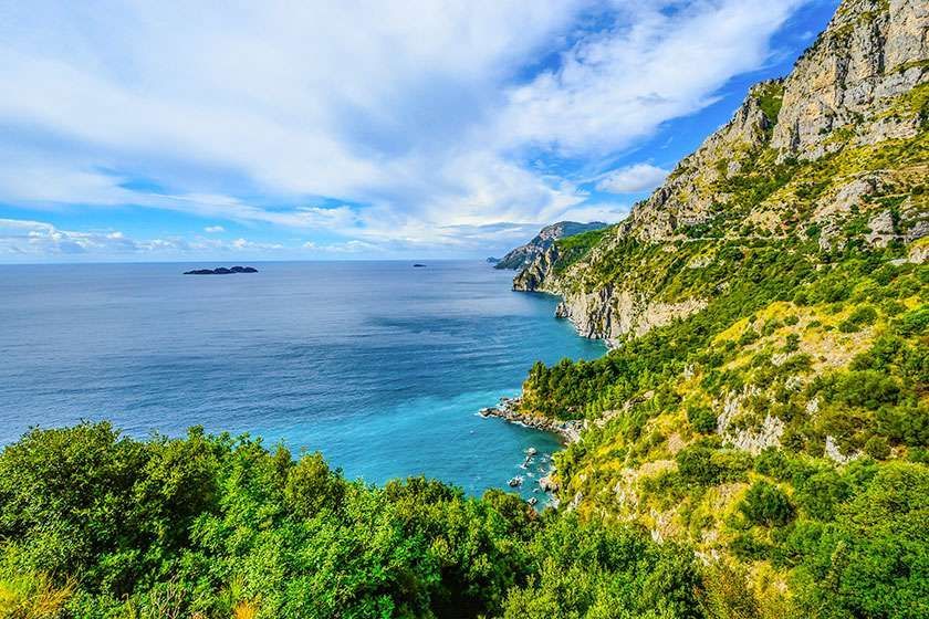 Lush green cliffs overlooking the turquoise Mediterranean Sea under a bright blue sky with light clouds.