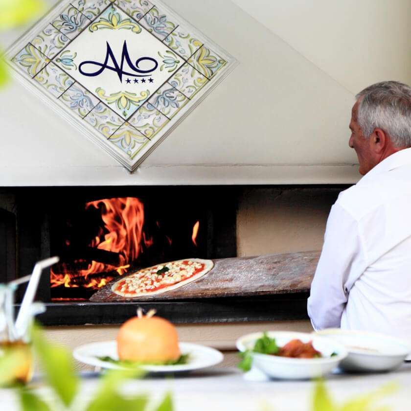 Una persona con una camicia bianca usa una pala di legno per infornare una pizza in un forno a legna, vicino ad altre pietanze già pronte.