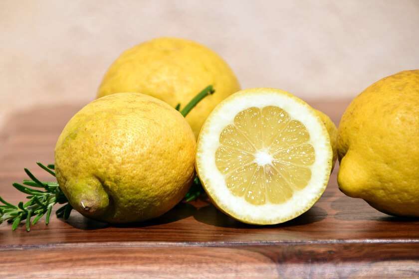 Diversi limoni di un giallo brillante, tra cui uno tagliato a metà, poggiano su una superficie di legno con un rametto di rosmarino verde.