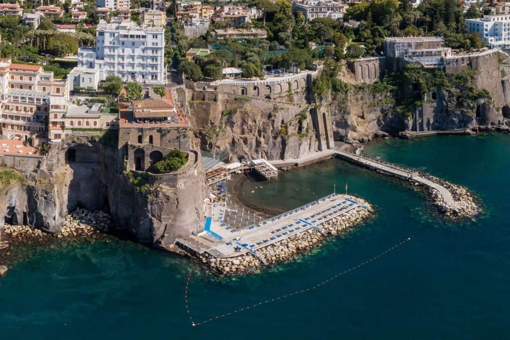 Aerial view of a Mediterranean coastal town with buildings atop cliffs, overlooking a small beach with rows of umbrellas.