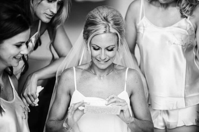 A bride in a veil reads a note, smiling, surrounded by three friends in satin pajamas in a black-and-white photo.
