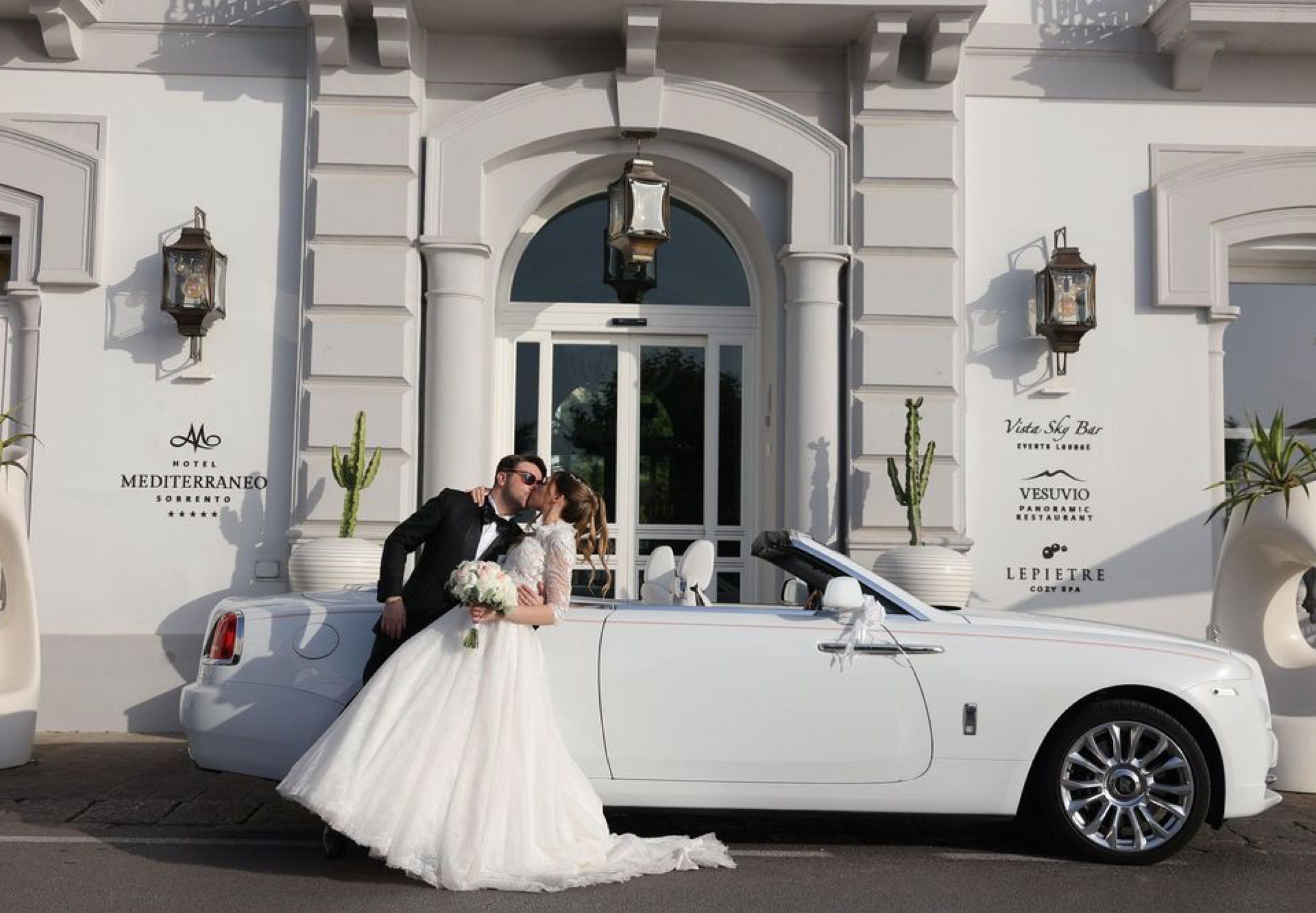 A couple in formal wedding attire kisses beside a white convertible in front of a white building with arched doorways.