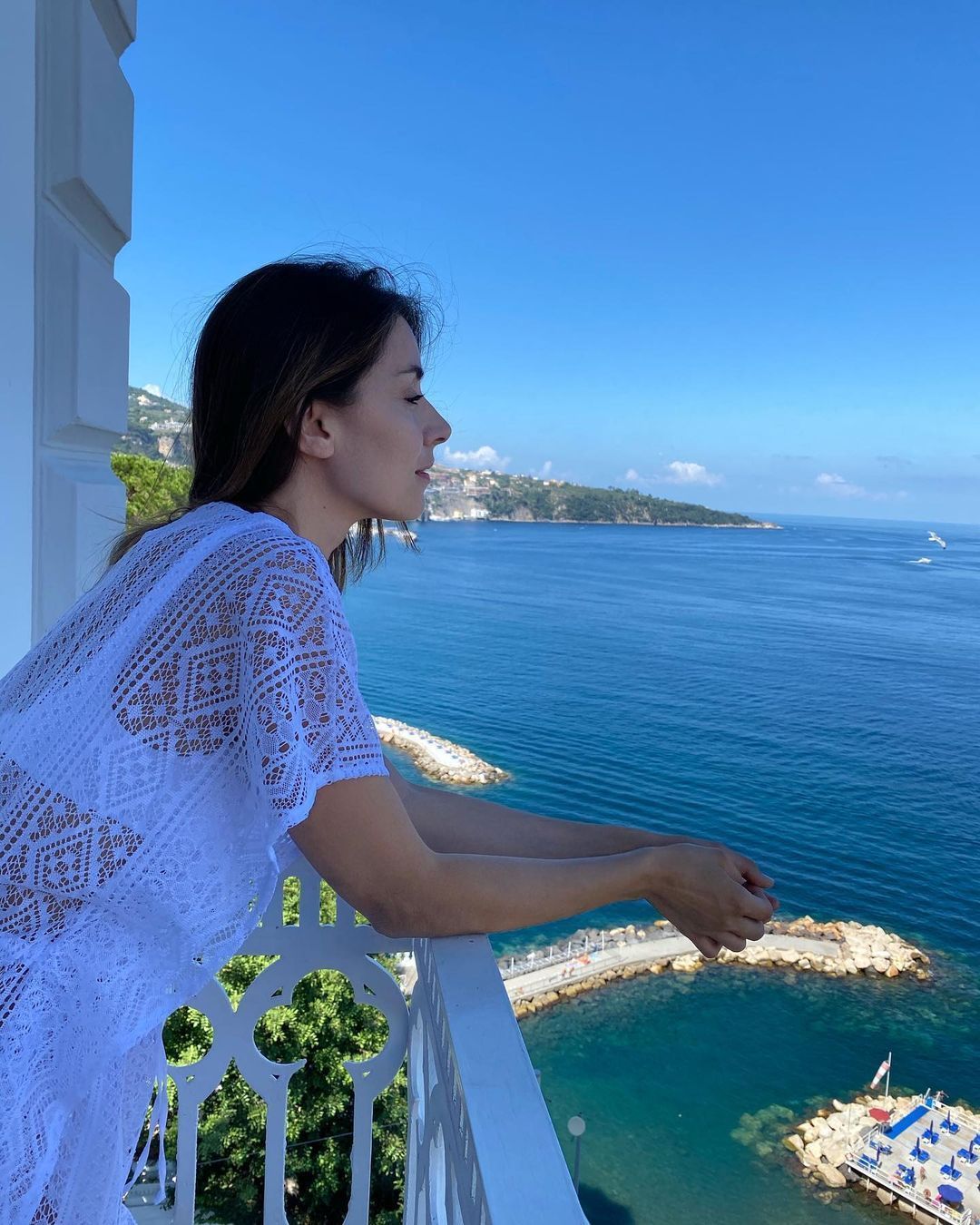A person in a white lace cover-up leans on a balcony railing, looking out at a blue sea and a rocky coastline.