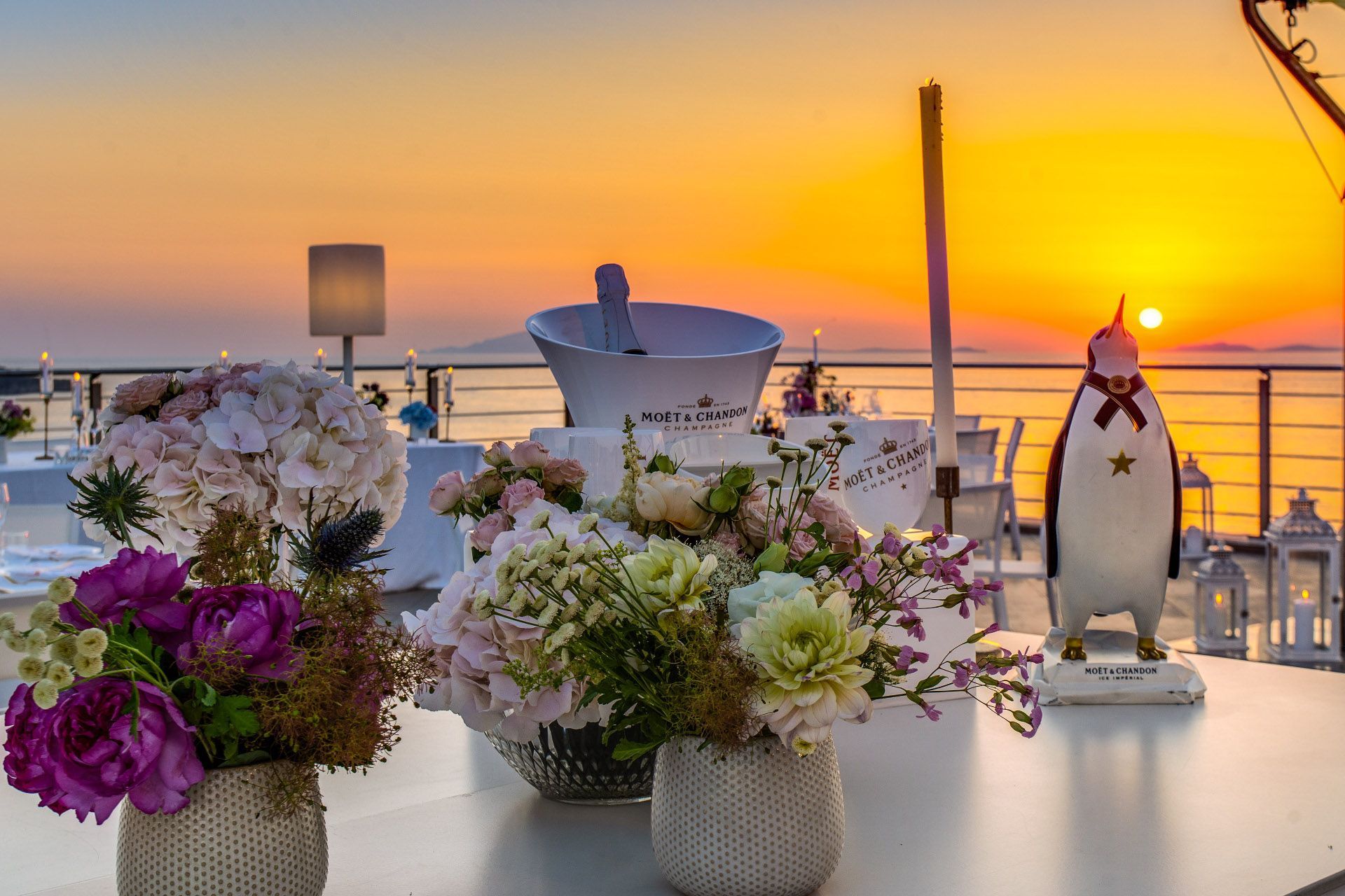 Un tavolo con composizioni floreali e un secchiello per lo champagne al tramonto, con una stravagante statuetta di pinguino, in riva al mare.
