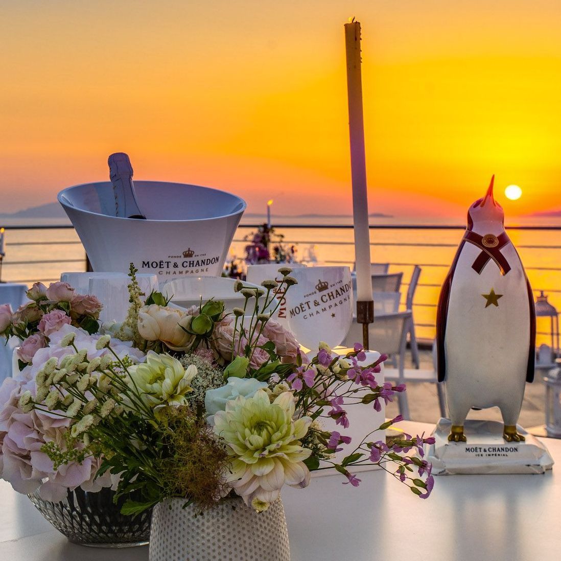 Una vista del tramonto con un secchiello per champagne Moët & Chandon, un centrotavola floreale e una statuetta decorativa di un pinguino su una terrazza.
