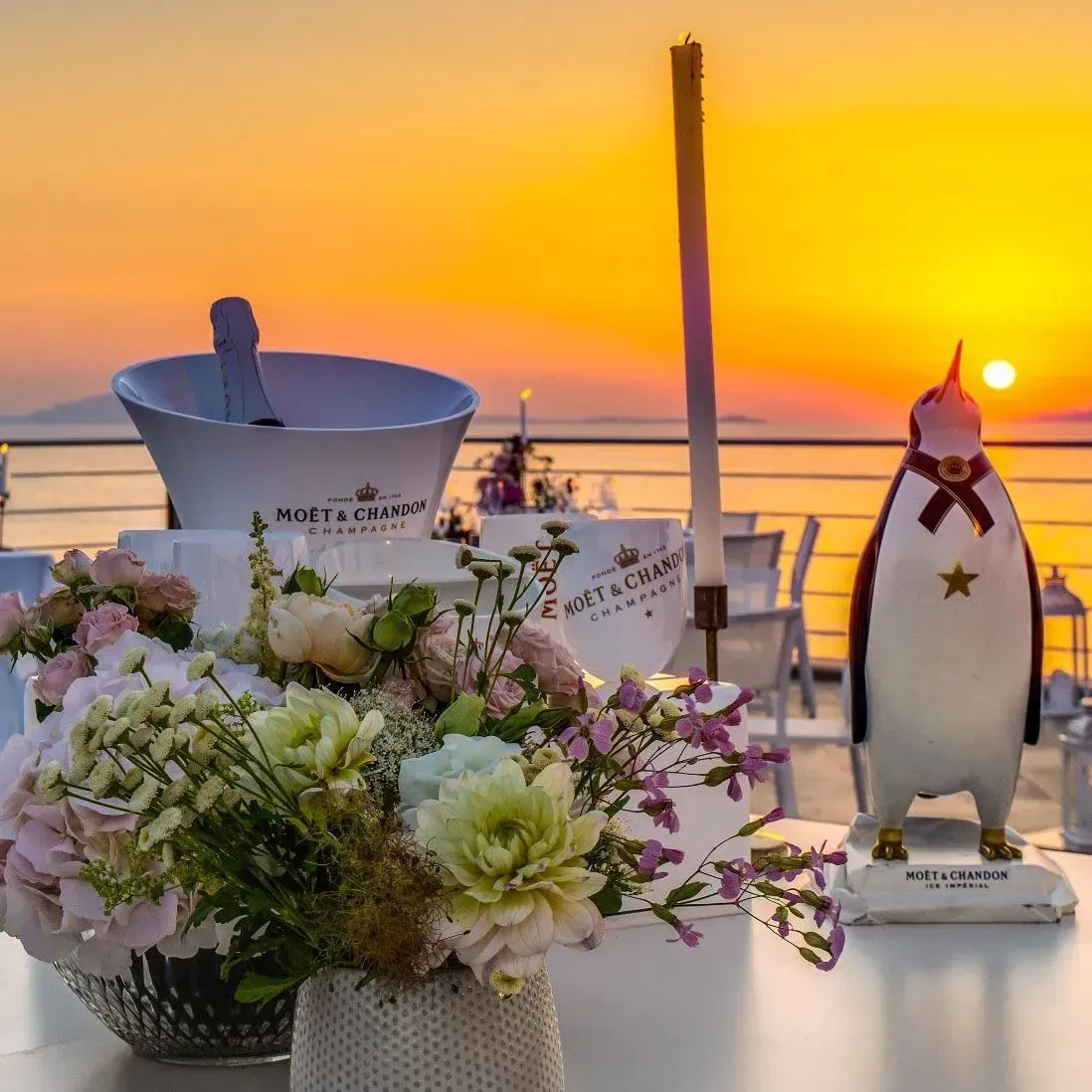 A bouquet of flowers sits on a table next to a Moët & Chandon ice bucket and a decorative penguin against a sunset sea.
