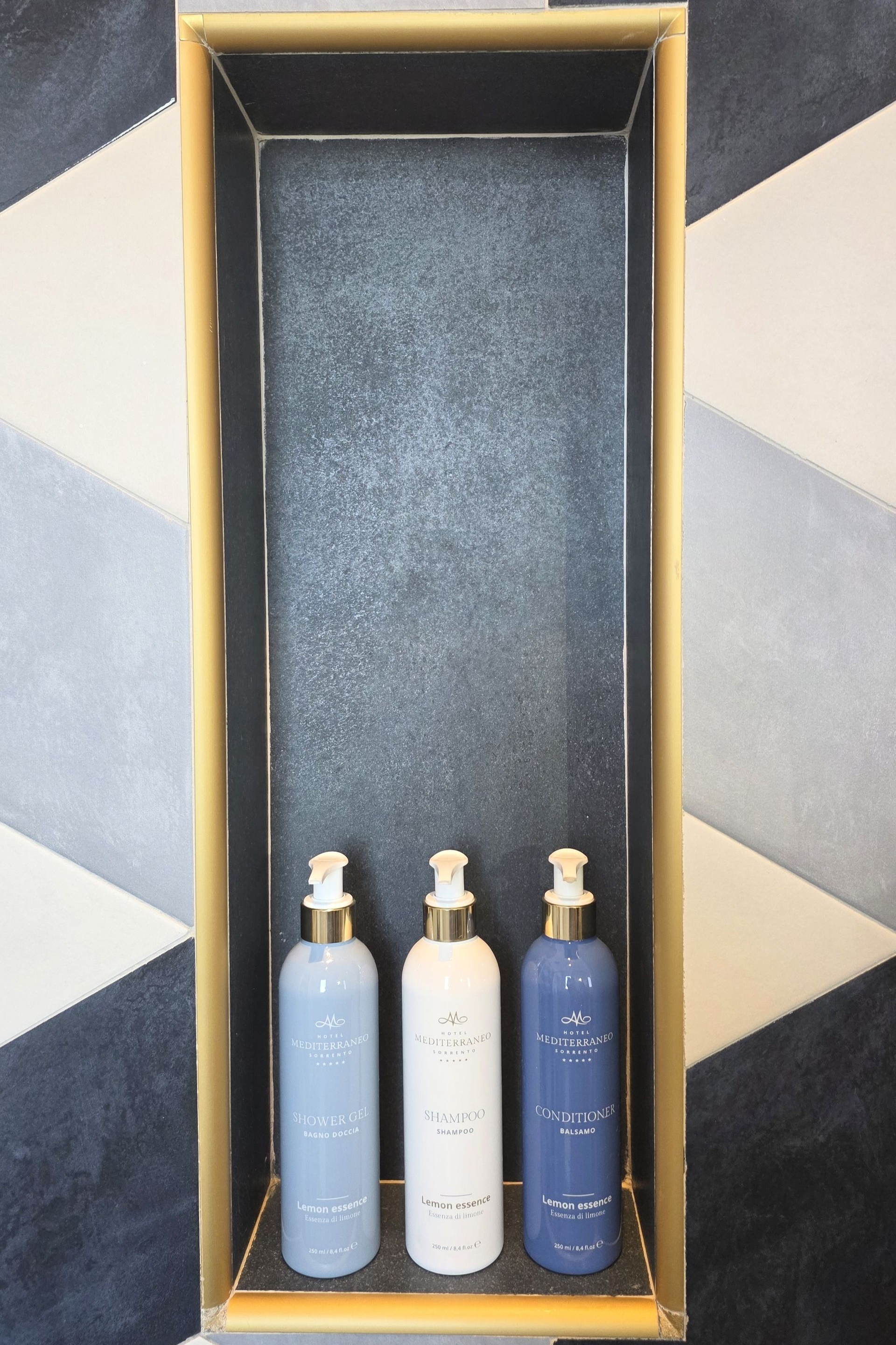 Tre flaconi con dosatore a pompa – uno azzurro, uno bianco e uno blu scuro – sono posizionati su un vassoio rettangolare alto e stretto con bordi dorati.