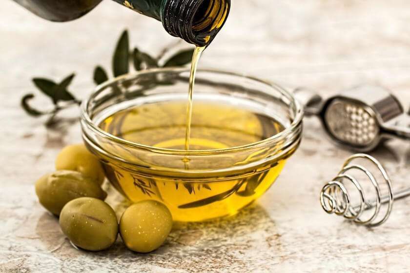 Olio d'oliva versato da una bottiglia in una piccola ciotola di vetro, circondata da olive verdi intere su una superficie di marmo.