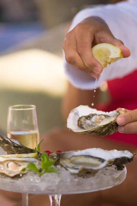 Una persona in abito bianco spreme del succo di limone su un'ostrica fresca adagiata su un letto di ghiaccio, con un bicchiere di vino sullo sfondo.