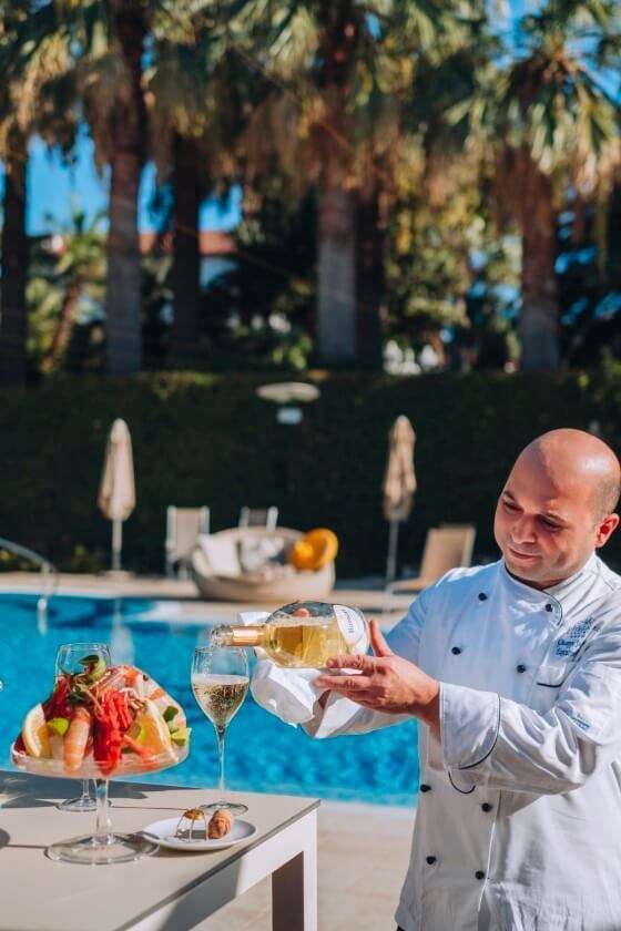 Uno chef versa del vino bianco in un bicchiere accanto a un piatto di frutti di mare a bordo piscina, con palme sullo sfondo.