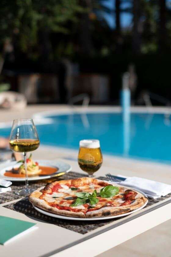 Una pizza, un bicchiere di vino e una birra serviti su un tavolo a bordo piscina, con una piscina di un blu brillante sullo sfondo.