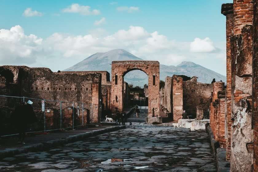 Antiche rovine in pietra di Pompei con una strada lastricata che conduce a un arco e il Vesuvio sullo sfondo.