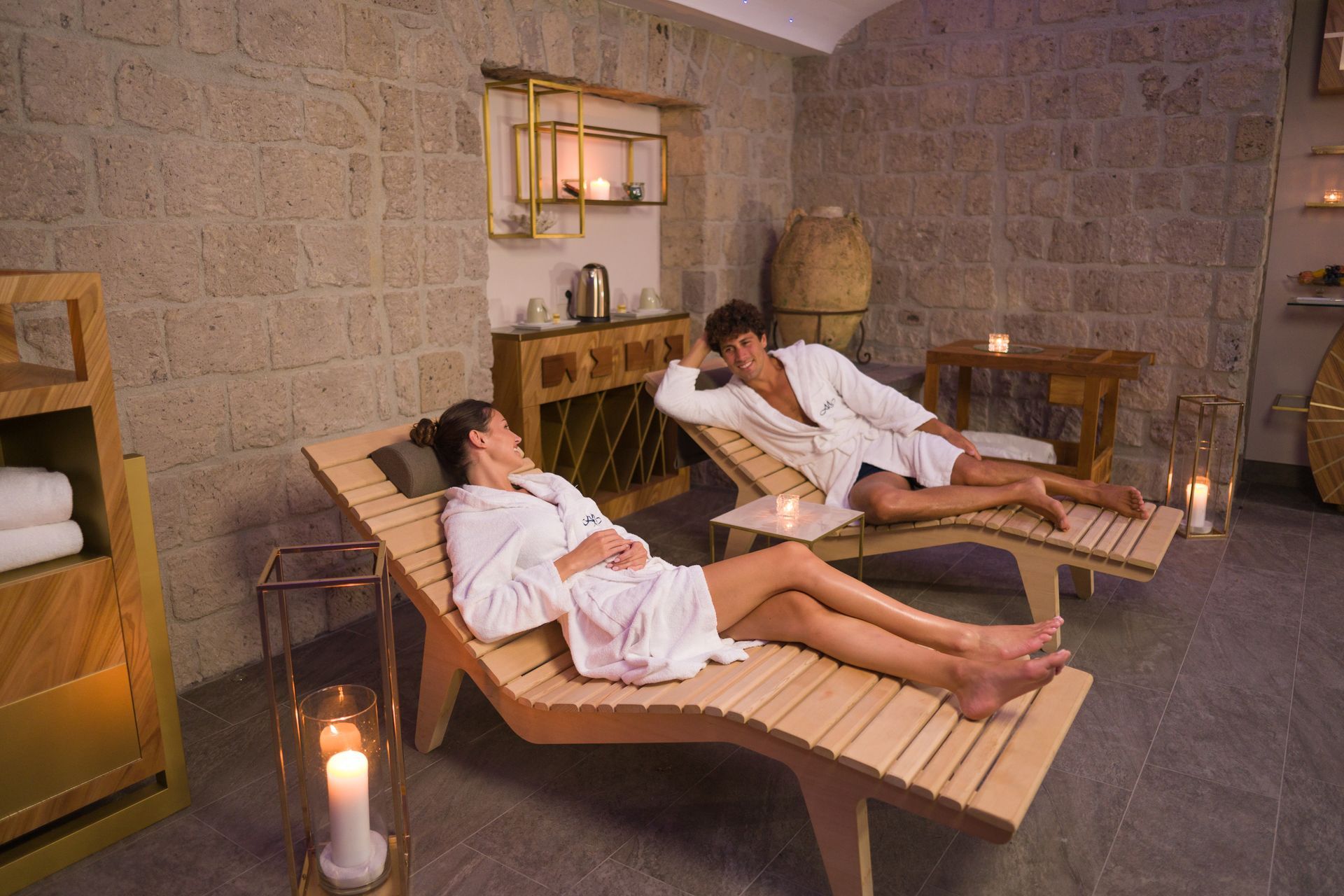 A man and a woman in white robes relax on wooden lounge chairs in a stone-walled spa room with soft candlelight.