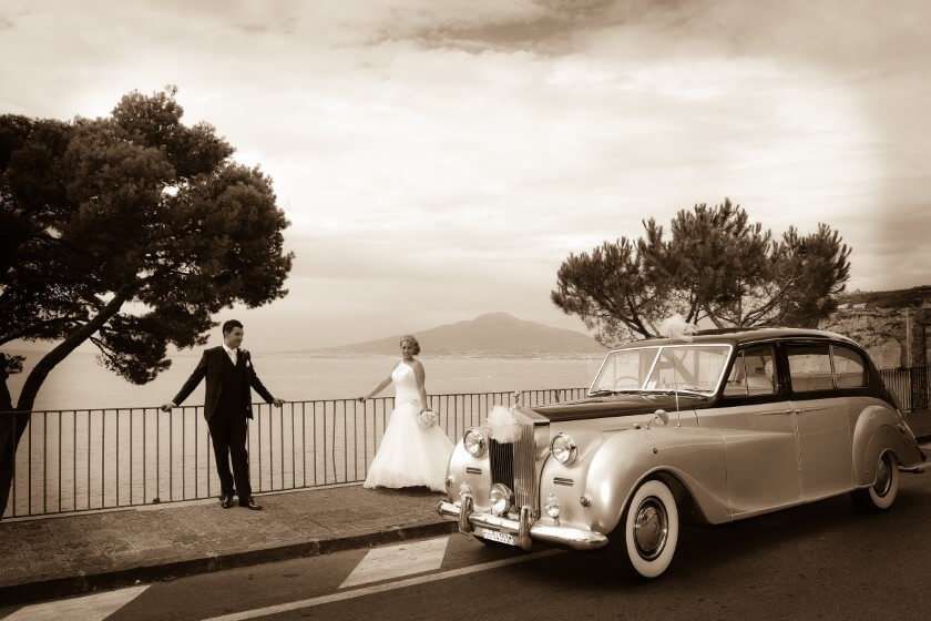 A bride and groom in vintage-style wedding attire pose near a classic car by a scenic coastal railing in sepia tones.