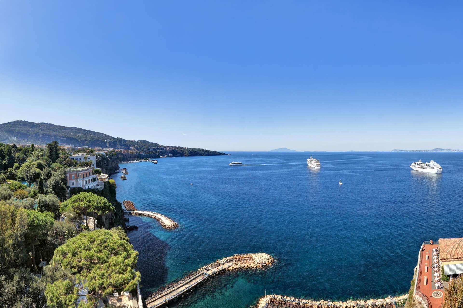 Vista panoramica di una costa mediterranea soleggiata con lussureggianti scogliere verdi, un molo in pietra e grandi navi da crociera in mare.