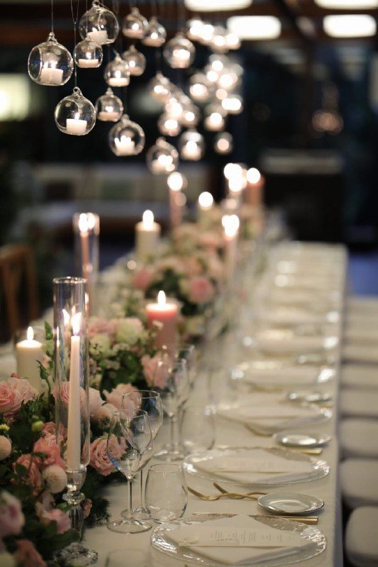 Long dining table set for an event with soft pink flowers, taper candles, and hanging glass orbs with tea lights above.