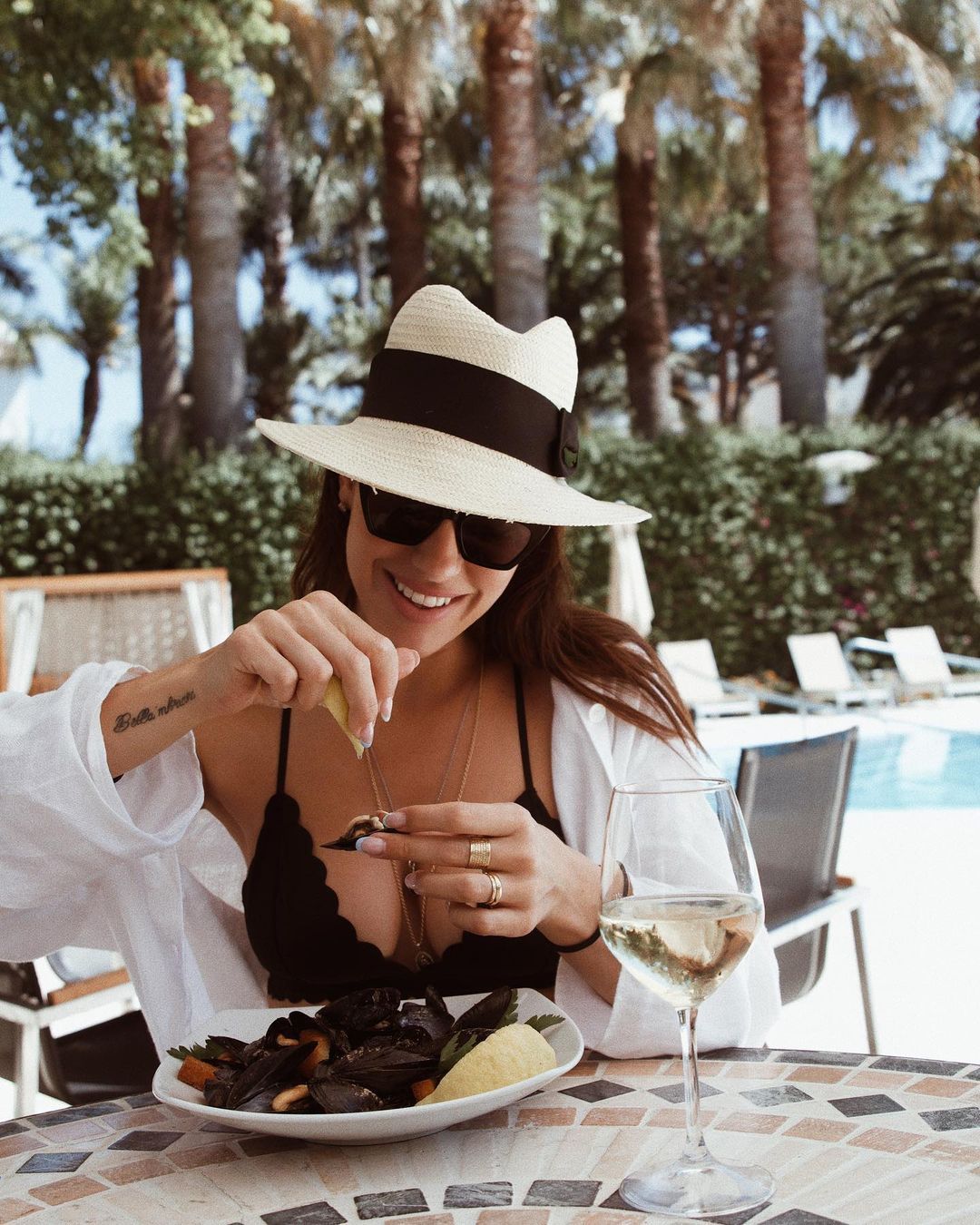 Una persona con cappello da sole e occhiali da sole sprema del limone su un piatto di cozze a un tavolo all'aperto a bordo piscina.