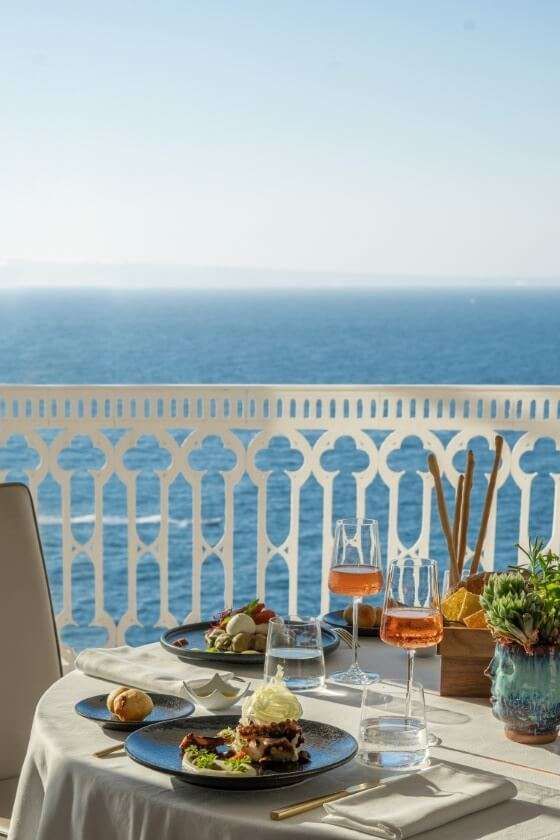Un tavolo per due apparecchiato per cenare su un balcone con vista su un vasto oceano blu, sotto un cielo limpido e luminoso.