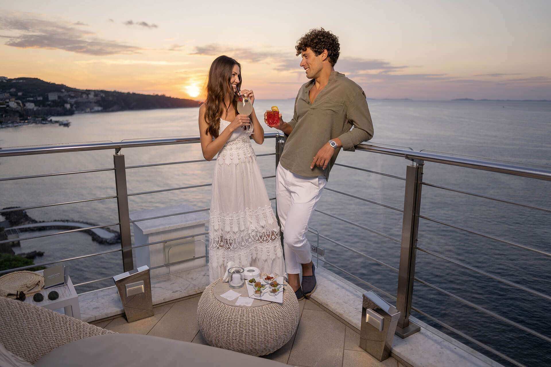 A couple holding cocktails on a balcony overlooking the ocean at sunset, dressed in white and olive tones.