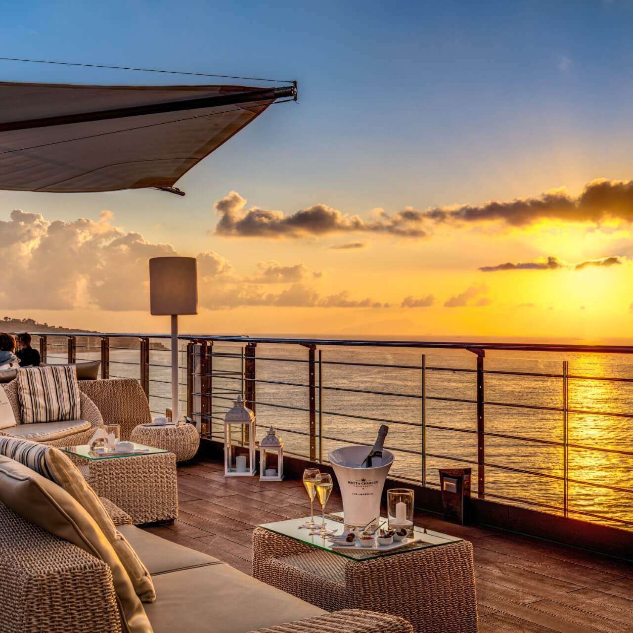A wicker sofa set on a balcony deck overlooking a golden sunset over the ocean, with a wine bucket on a side table.