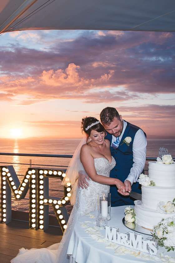 A couple in formal wedding attire cuts a multi-tiered white cake on a deck at sunset, with illuminated letters nearby.