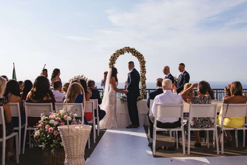 A couple stands under a floral arch during an outdoor wedding ceremony, with guests seated in rows facing them.