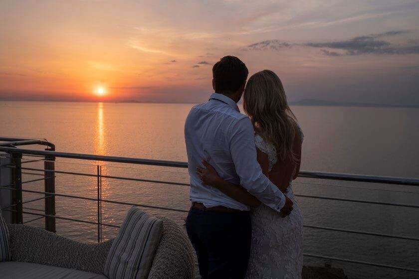 A couple in wedding attire stands on a balcony, embracing while overlooking the ocean during a golden sunset.