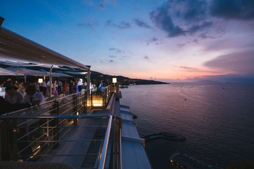 People gathered on an outdoor restaurant terrace overlooking the ocean during a vibrant sunset.