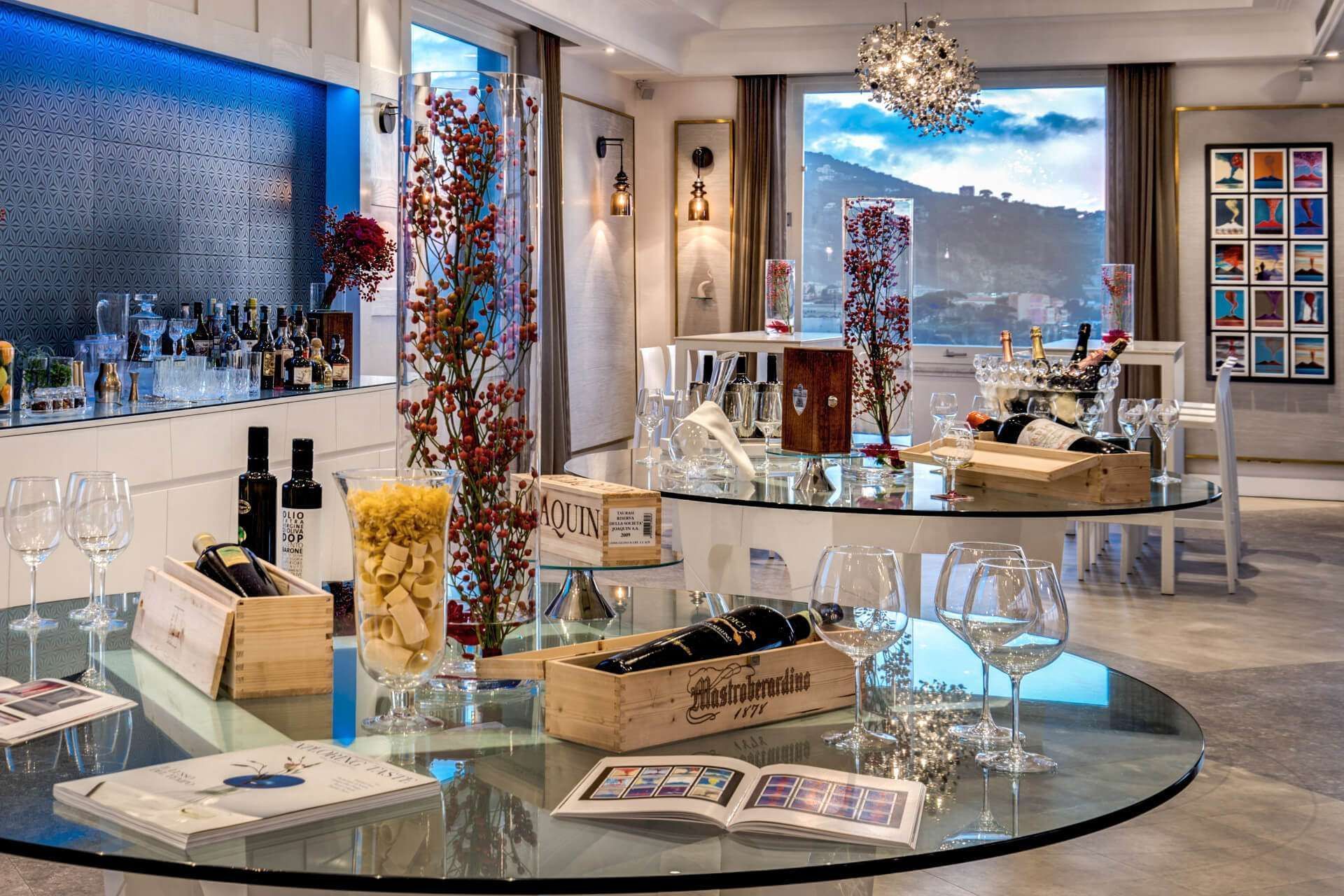 Un salone moderno con tavolini in vetro che espongono bottiglie di vino, bicchieri e piante decorative, il tutto sullo sfondo di un paesaggio suggestivo.