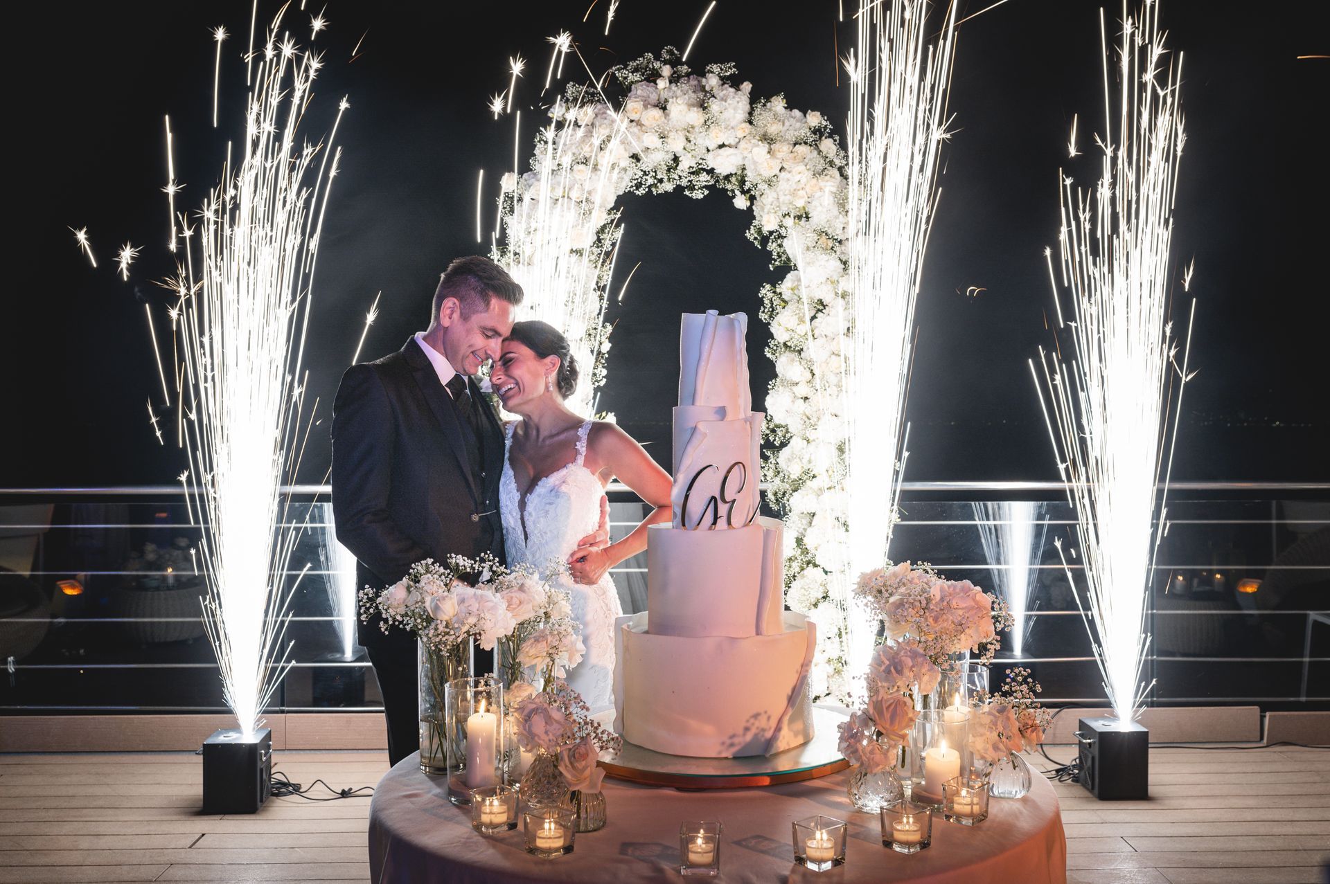 A couple stands embracing beside a tiered wedding cake at night, framed by a floral arch and illuminated by fireworks.