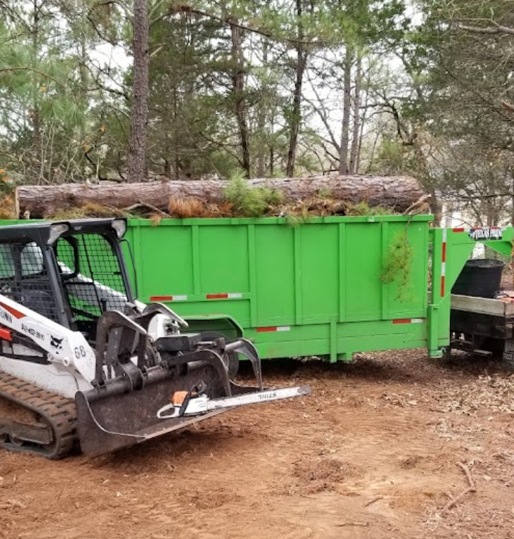 A green dumpster is being pulled by a bulldozer.