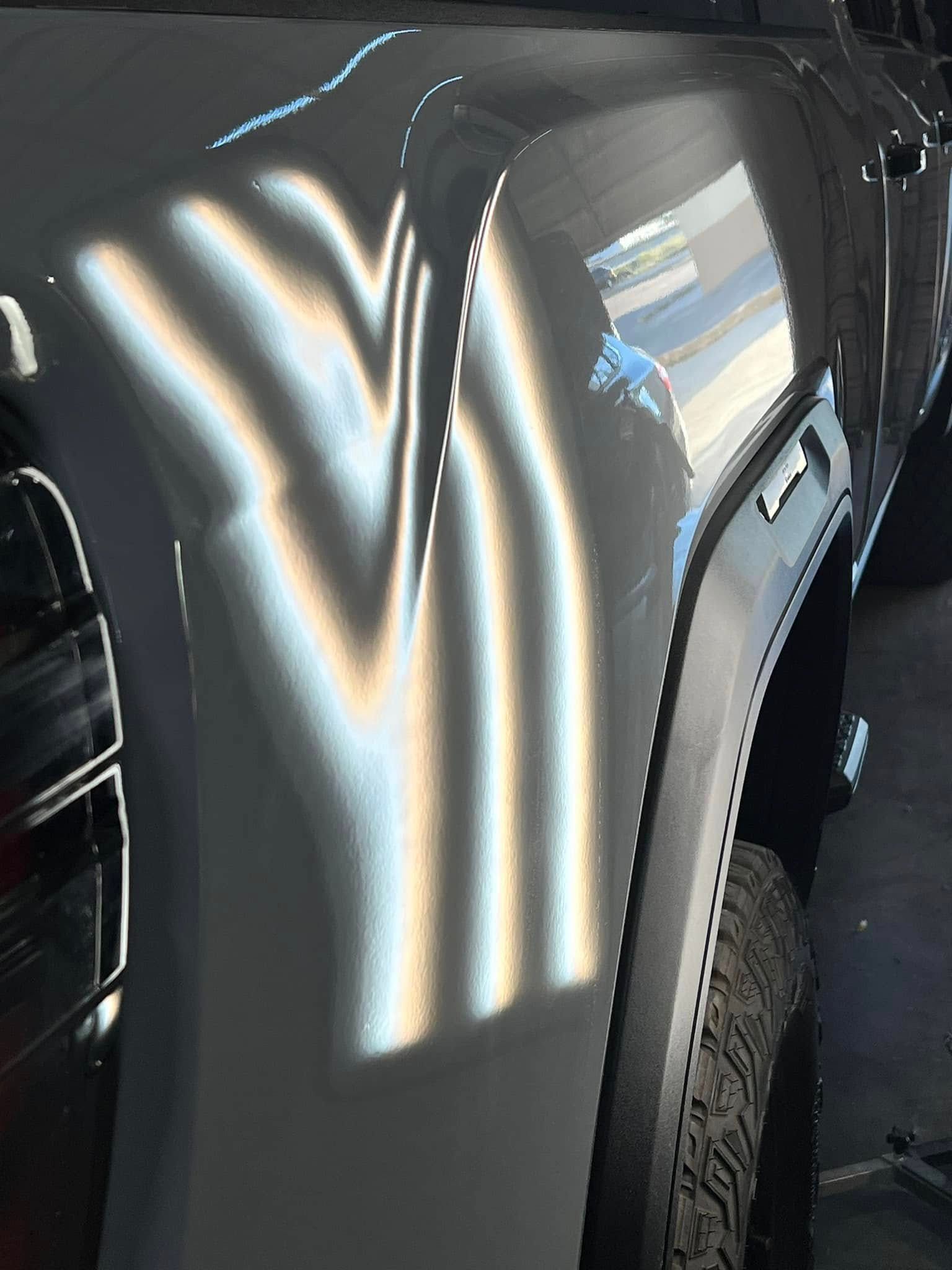 A dented gray car panel with reflection patterns, revealing damage near the rear tire.