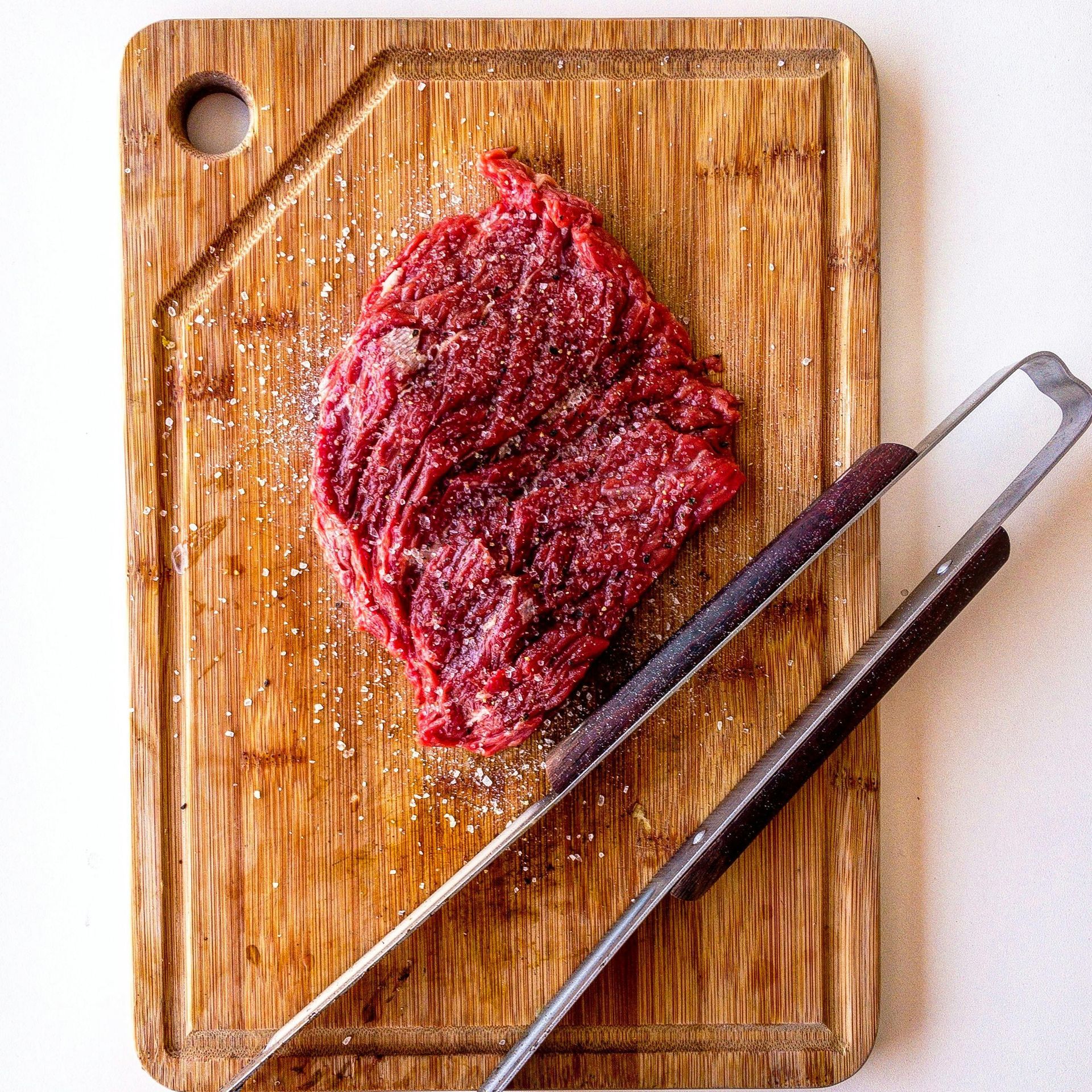 Raw steak seasoned with salt on a wooden cutting board, tongs nearby.