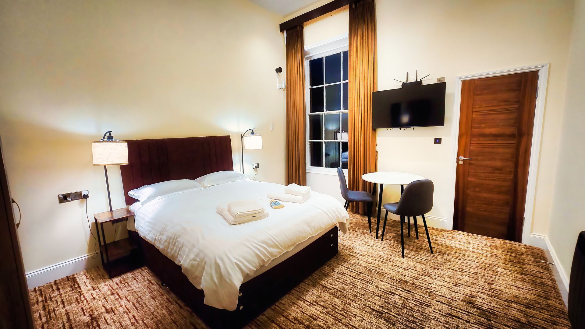 Hotel room with bed, window, TV, table, and door. Warm lighting. Carpeted floor.
