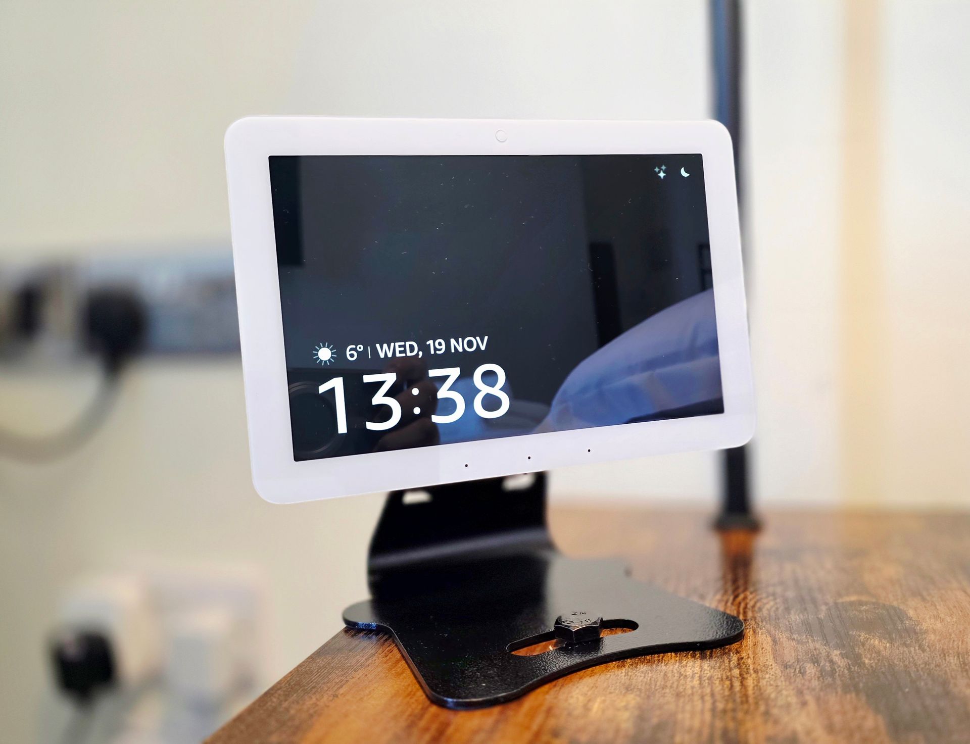 White tablet on black stand displaying time, date, and weather info, resting on a wooden surface.