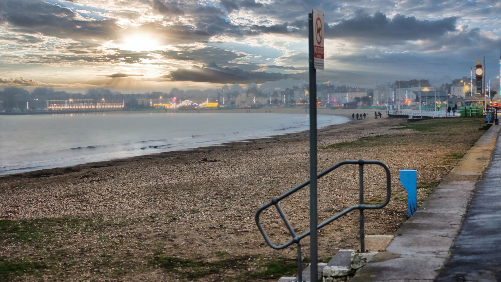 Beach scene at dusk with calm water, cloudy sky, and distant lights. Sign and handrail in foreground.