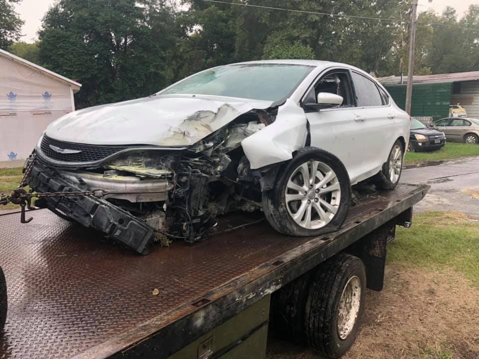 White car with severe front-end damage, on a tow truck, outdoors.