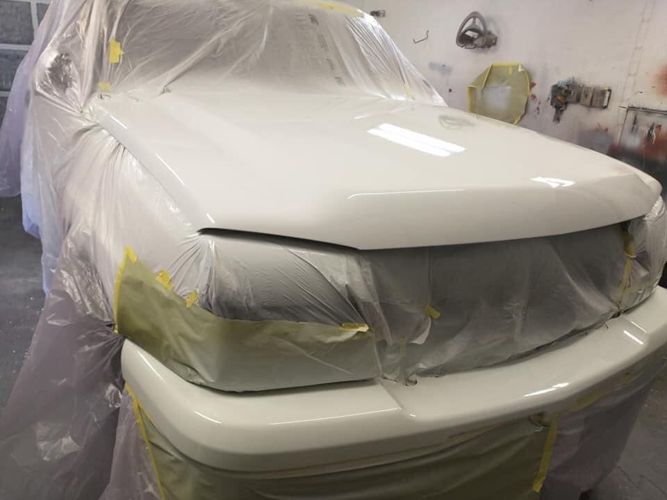 White car hood and bumper freshly painted, covered in plastic and tape in a workshop.