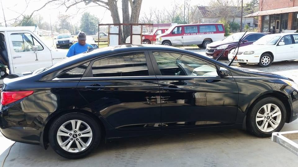 Black sedan parked outside; person in blue shirt nearby. Other vehicles and buildings in background.