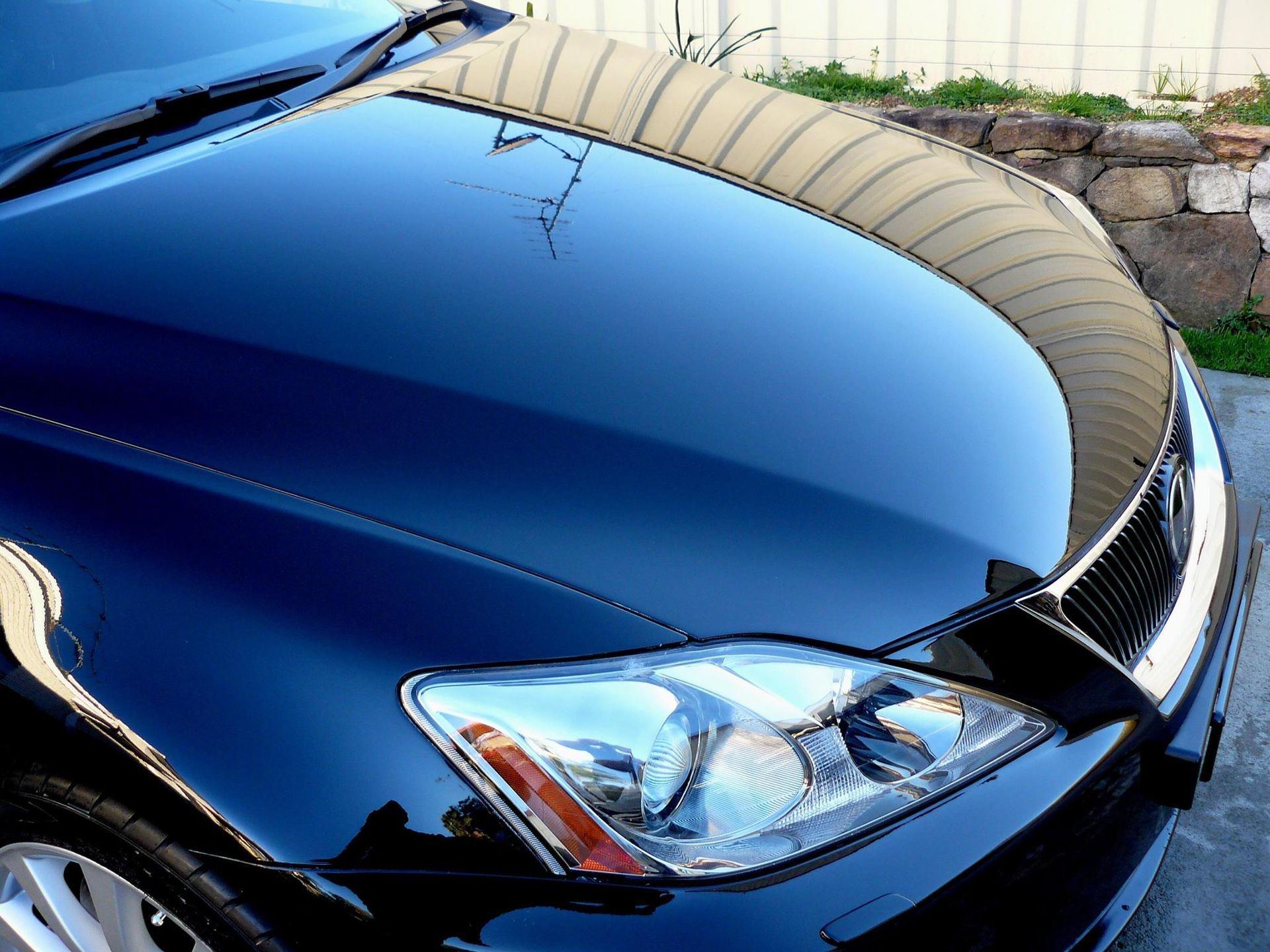 Black car hood reflecting sunlight, chrome headlight, Lexus emblem visible.