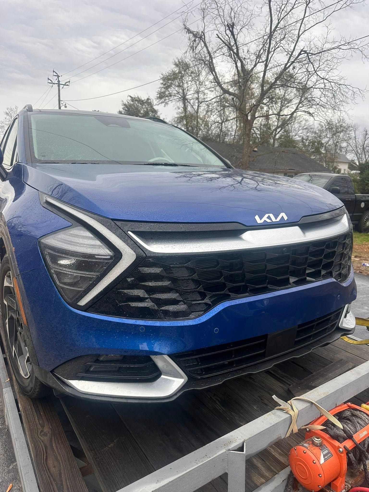 Blue Kia SUV on a trailer, front view. Cloudy sky, trees in the background.