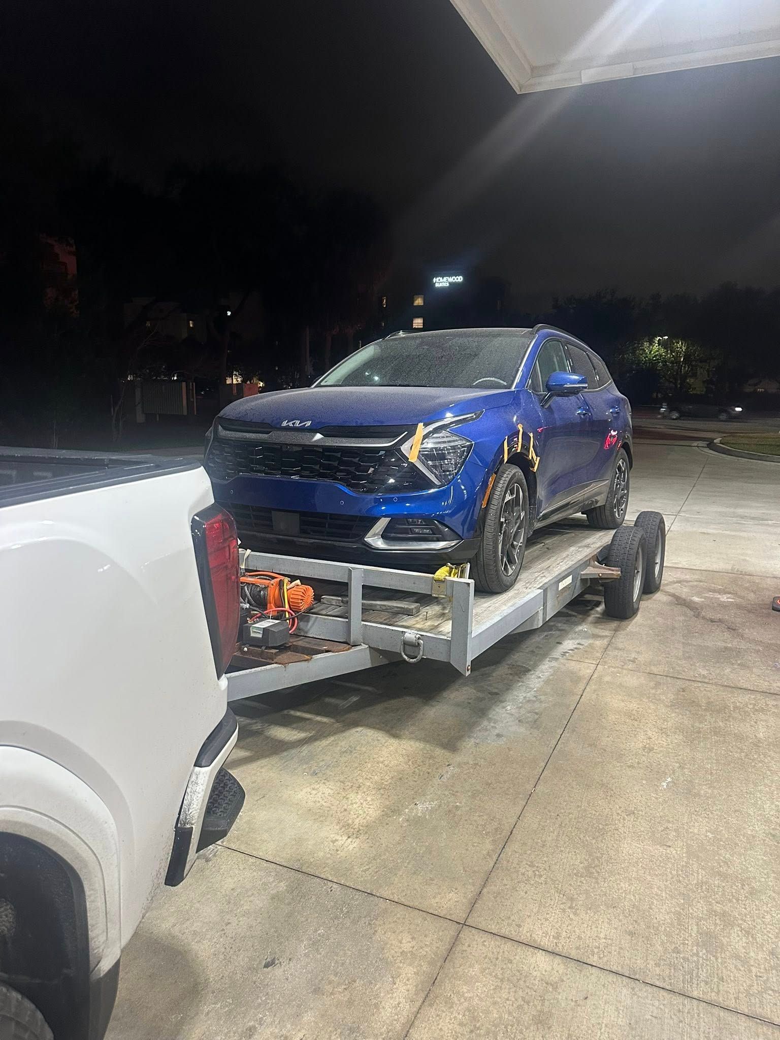 Blue car on a flatbed trailer being towed by a white truck at night.