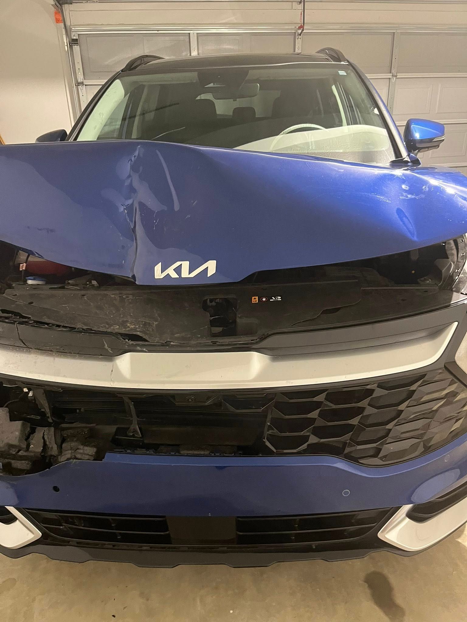 Damaged blue Kia SUV in a garage, front end caved in.