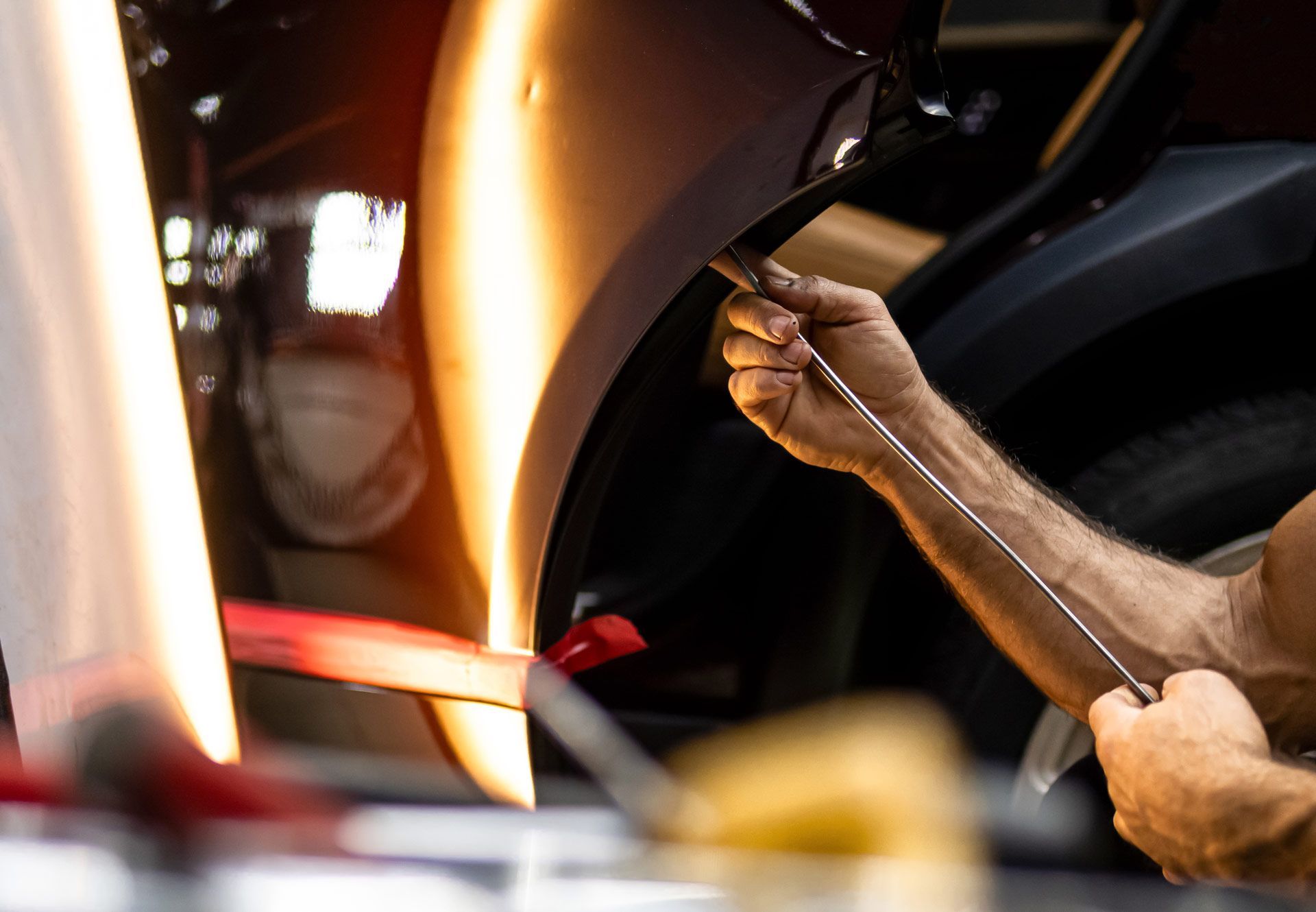 Person uses a tool to repair a dent on a car's side panel, with reflective light and repair tools visible.