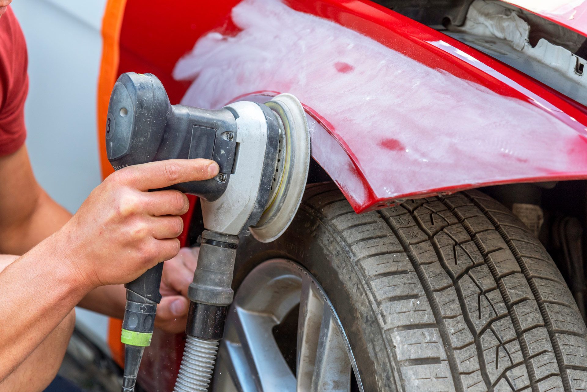Person sanding a red car fender with an electric sander, near a car tire.