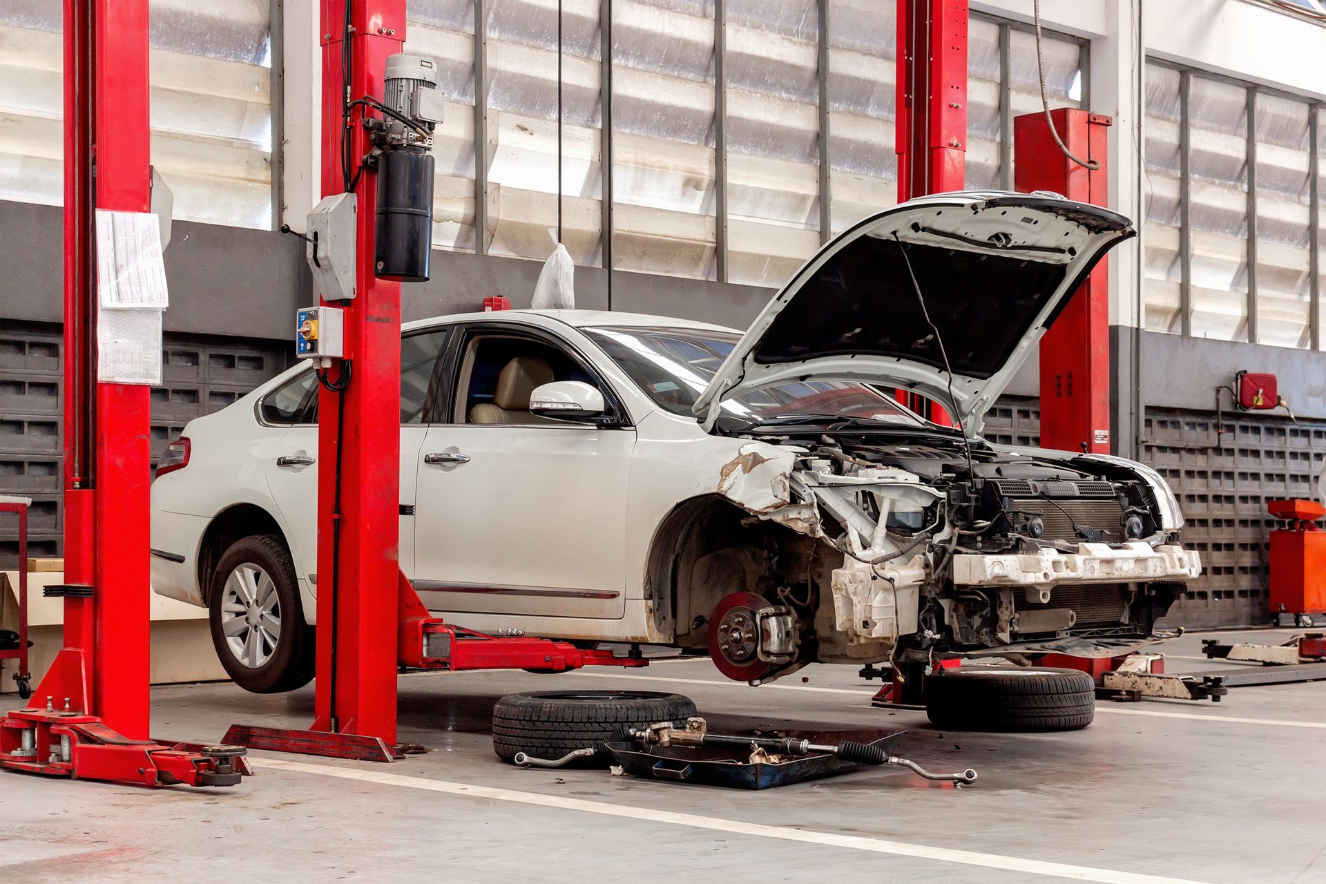 White car on a lift in a repair shop with its hood open and front bumper removed.